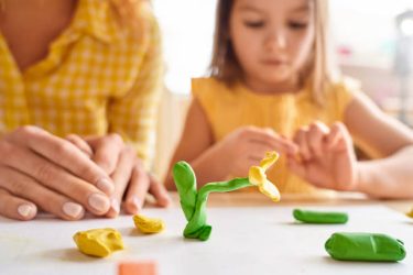 Selective focus on plasticine flower against blurred background with young mother and her junior daughter spending day together, playing at home