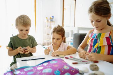 Three young children engaged in creative art activities at a table with markers, colored pencils, and other art supplies.